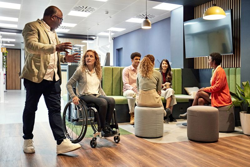 Full length view of small group sitting together and talking as mature man and woman walk and wheel past, face to face and exchanging ideas.