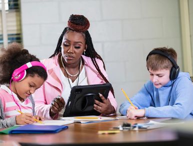 Teacher helping two students with their schoolwork, one with pink headphones and the other with black headphones.