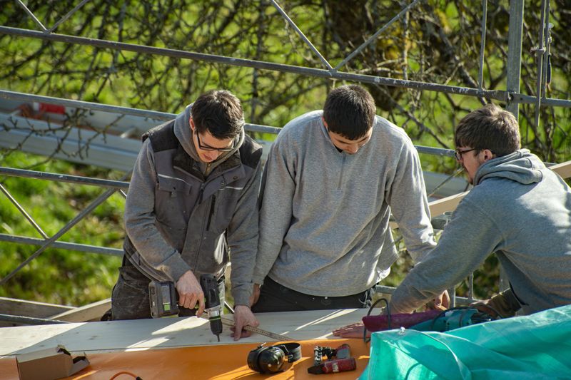Three young carpenters working on a roof. A carpenter with impact driver screws a screw into a wooden board. Apprentice learns from trainer.