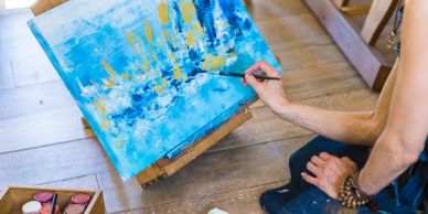 Person painting an abstract blue and yellow artwork on a canvas on the floor.