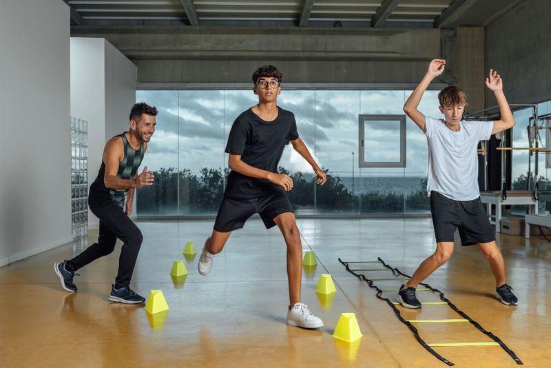 Teenagers and instructor performing circuit routine functional training at the gym. Male coach and Two boys jumping squats in a big gym with windows. Horizontal