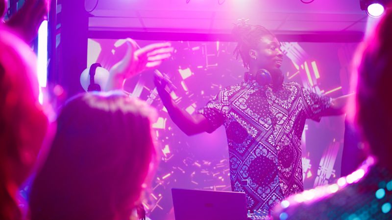 Male DJ using audio mixing station at party, putting modern music for crowd of people on dance floor. Young persons having fun partying with stage lights and live performance. Handheld shot.