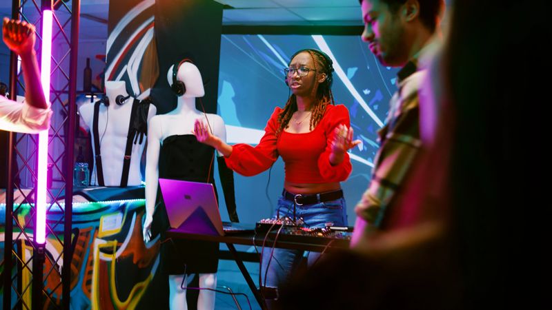 Funky DJ using mixing station on stage to mix music at party, having fun with diverse people in discotheque. Cheerful cool woman partying and using audio stereo panel at club. Handheld shot.