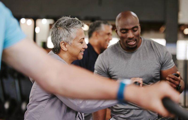Senior group, exercise bike and personal trainer for fitness, health and retirement wellness by blurred background. Elderly woman, bicycle training and diversity with black man, advice and motivation