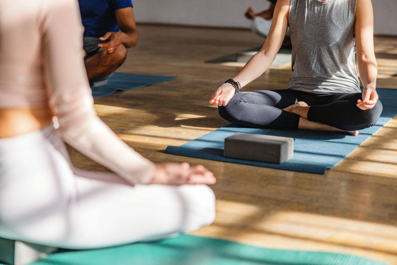 Diverse yoga class participants sitting on blue and green yoga mats in a Sukhasana pose during their yoga session at the beautiful studio with wooden floors and natural light. The sunlight is creating beautiful patterns on the wooden floors.