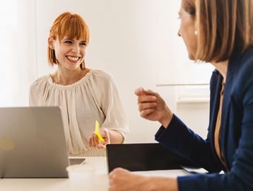 Two women engaged in a friendly office discussion with laptops.