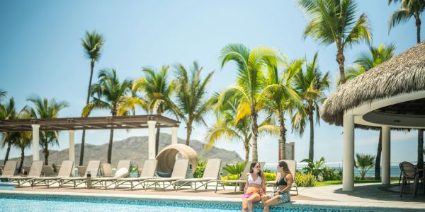 Dos mujeres conversan junto a una piscina en un día soleado.
