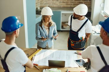 Construction team discussing blueprints on site wearing safety helmets. Our Services include: Home office cleaning, Landscaping services and Tree care services on power washing services and handyman services.