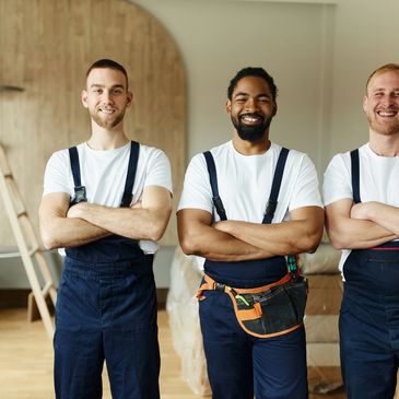 Three smiling workers in overalls with arms crossed standing indoors.