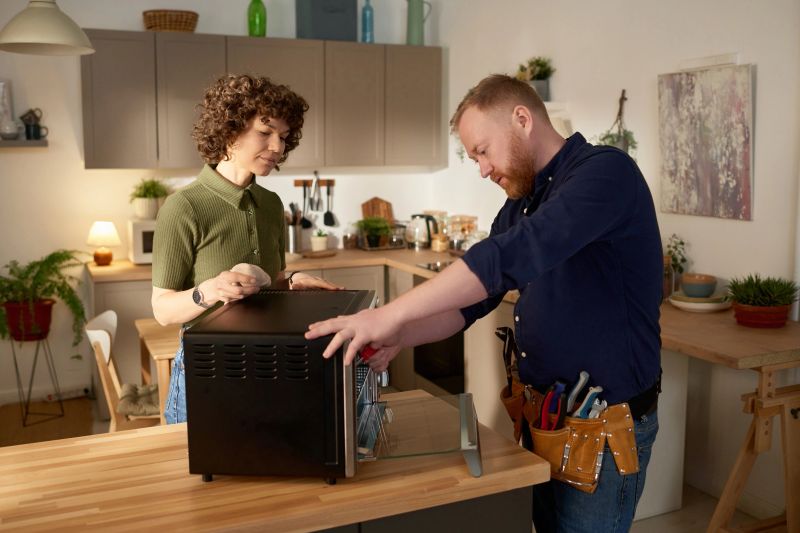 Repairman using his tools to fix microwave oven while standing in the kitchen together with housewife