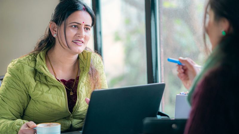 An Asian/Indian mid-adult, confident, and happy businesswoman sits with a laptop in a meeting with her female client. She drinks coffee with her during the discussion in the daytime office.