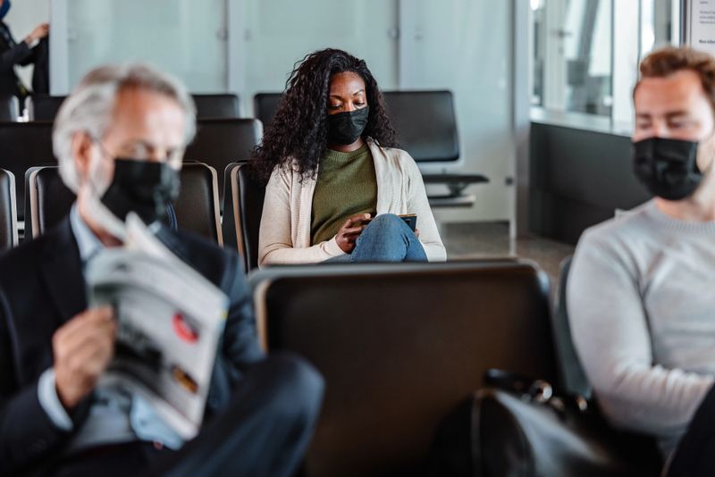 Medium side shot with blurred sides of a multiracial group of people waiting for their flight at the airport gate while wearing masks and trying to be busy in different activities like browsing and reading to not get bored.