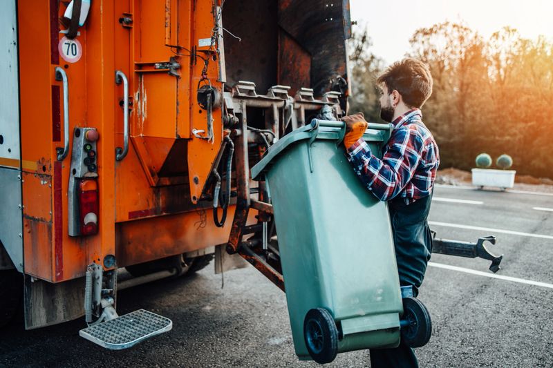 Young garbage man working on city street and emptying dustbins for trash removal. Bright sunny day.