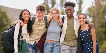 Five diverse teenagers smiling outdoors with backpacks, showing friendship.