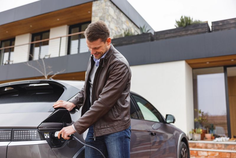 A smiling adult Caucasian male charging his expensive electric vehicle in front of a luxurious residential building outside the city. He looks happy. Future of sustainable transportation and zero-emission mobility.