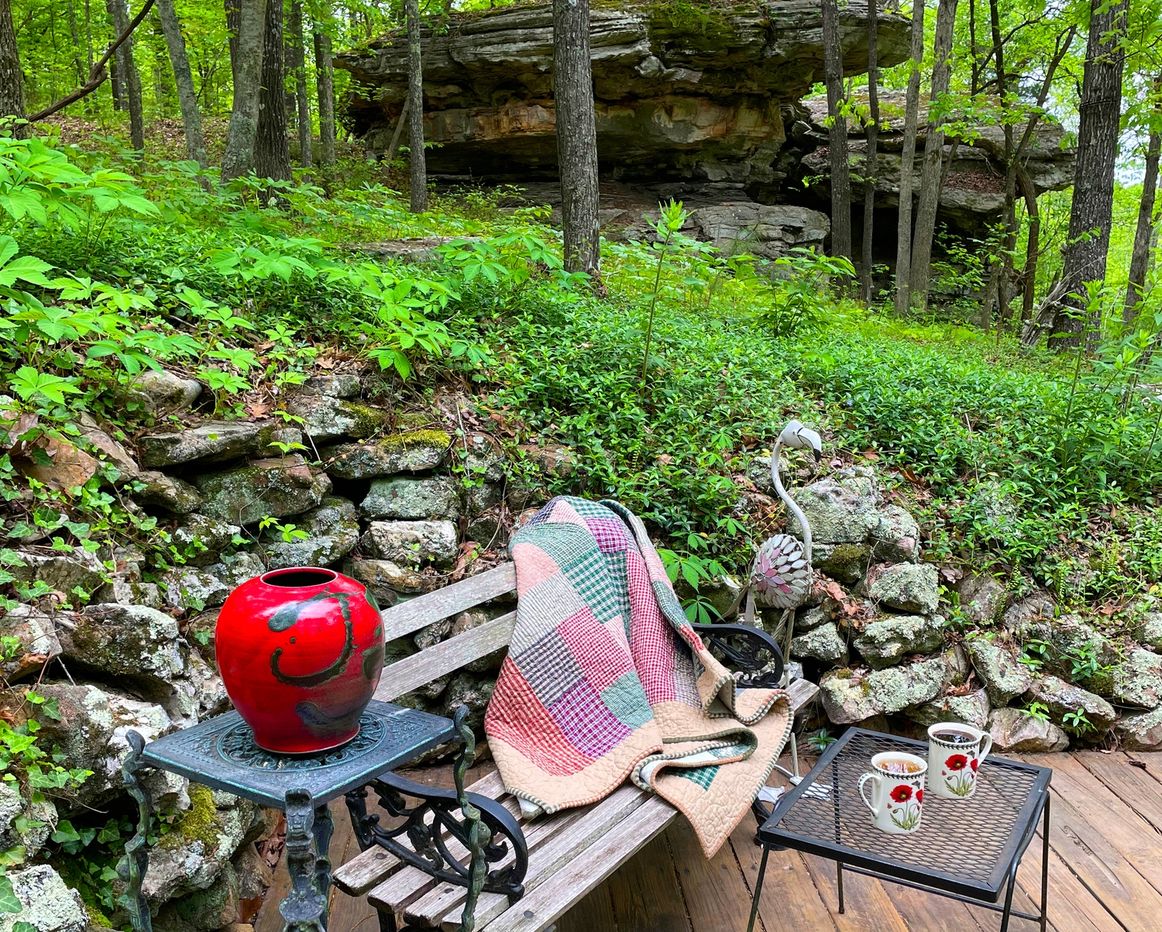 Cozy outdoor seating area with a quilt and two coffee mugs in a lush green forest.