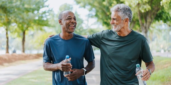 Two men walking outdoors, smiling and holding water bottles.
