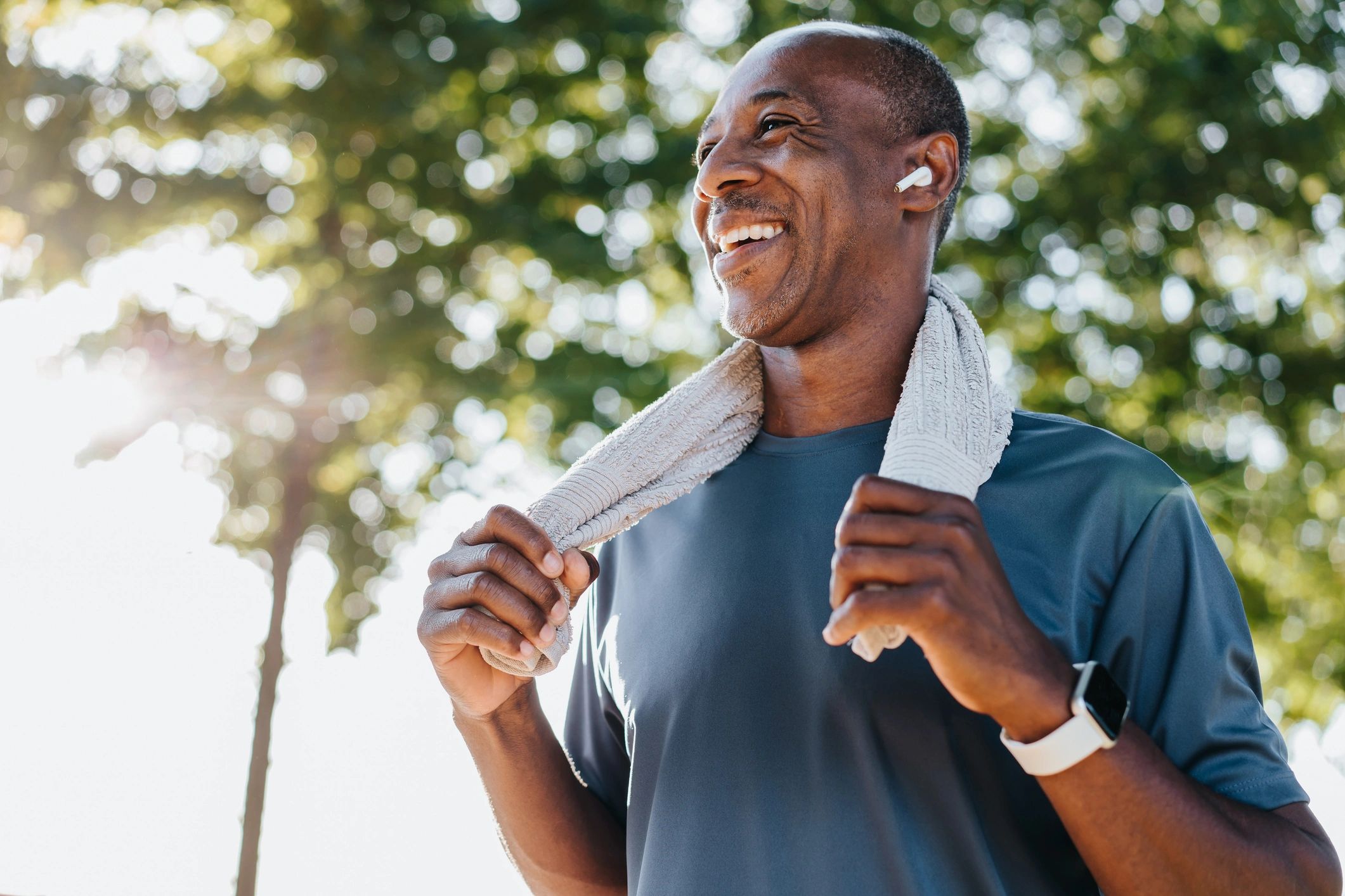 Smiling man with towel and earbuds outdoors in sunlight.