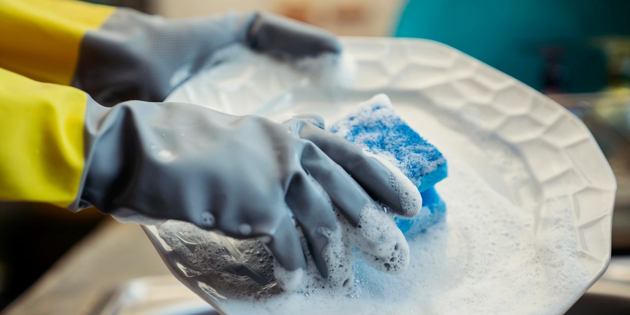 Closeup of cleaning gloves and sponge scrubbing white plate with soap during dish cleaning service