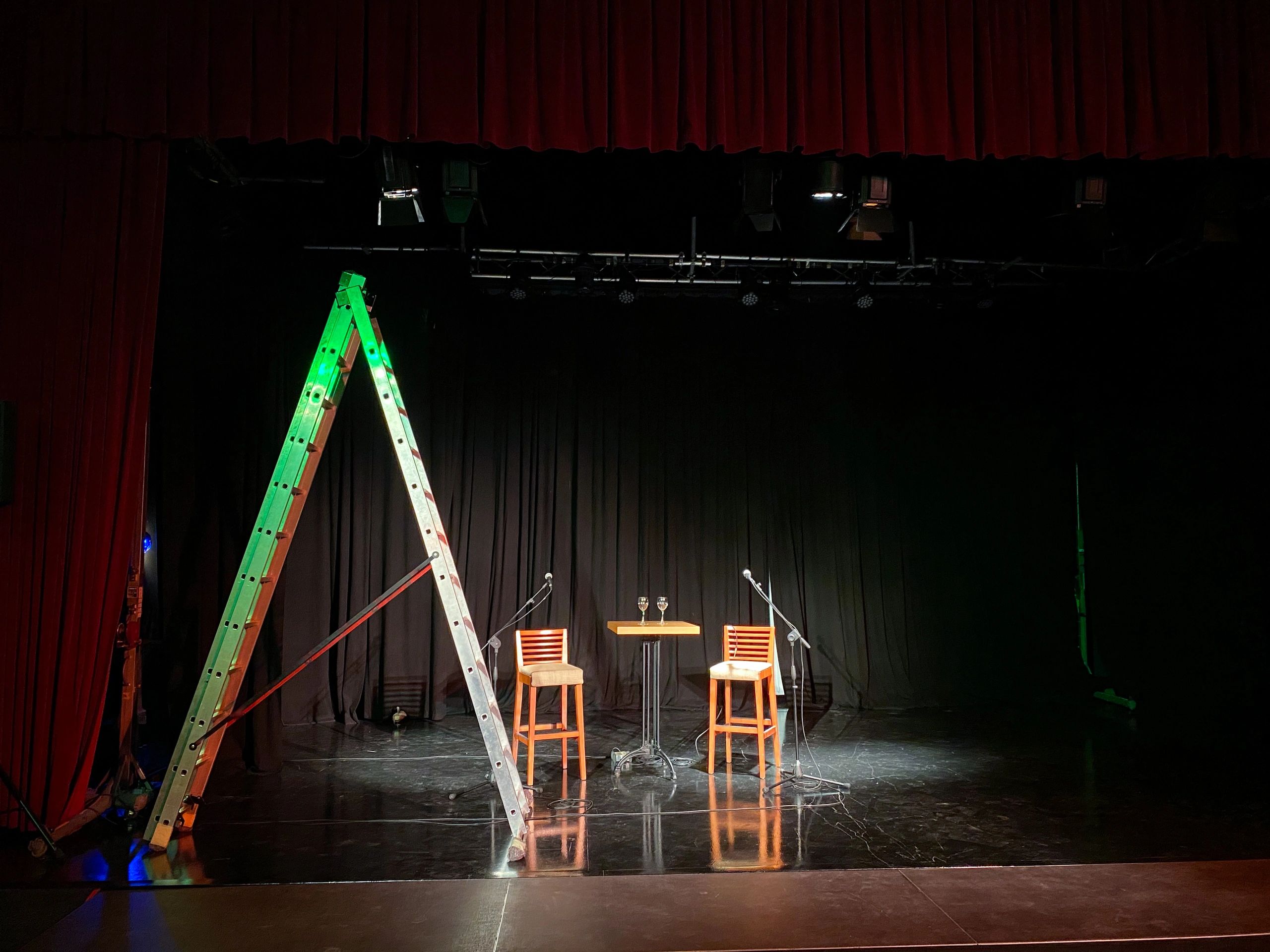 Two chairs and a table with glasses on a dimly lit stage with a ladder.
