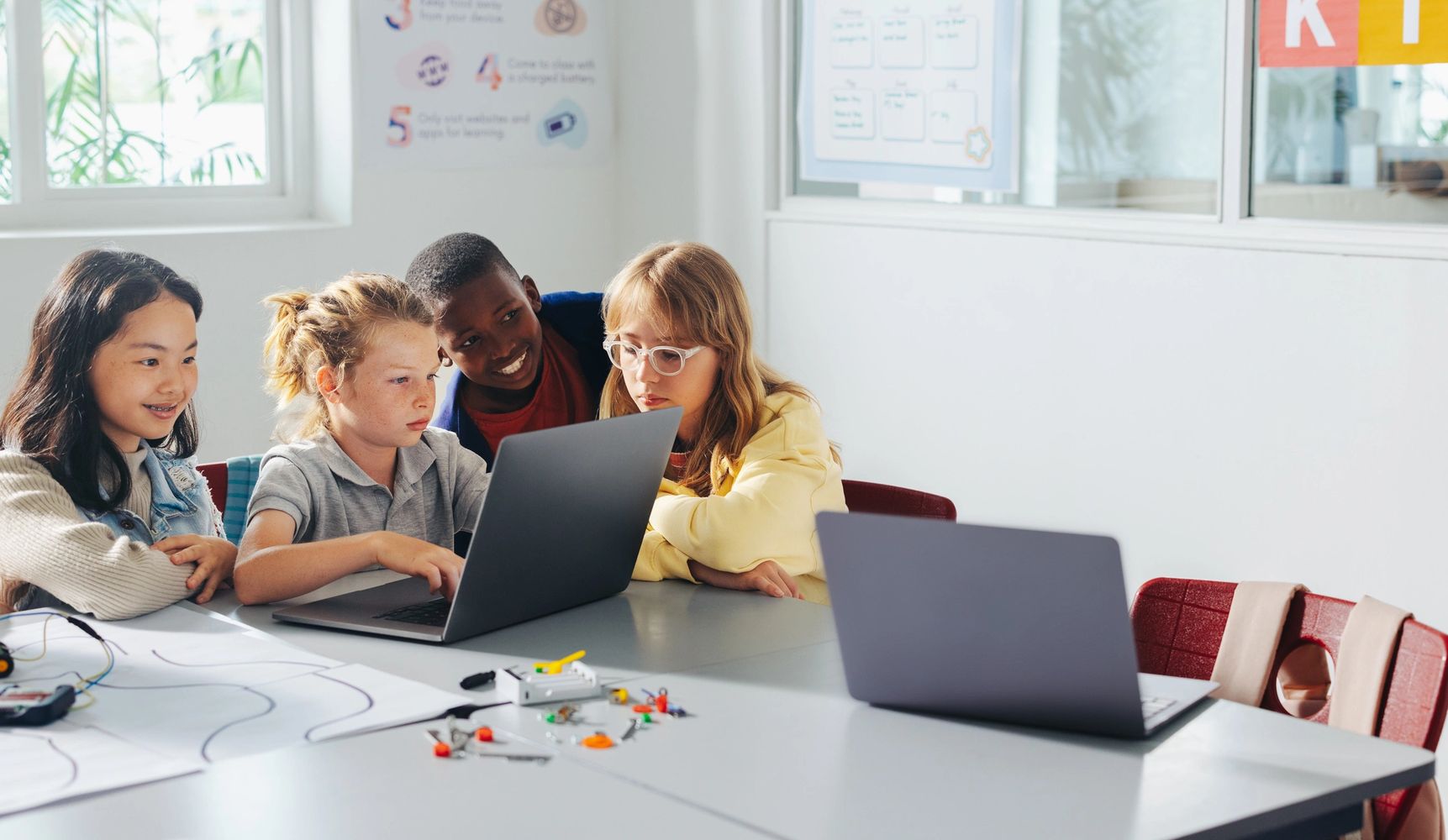 Four children collaborating around a laptop in a bright classroom setting.