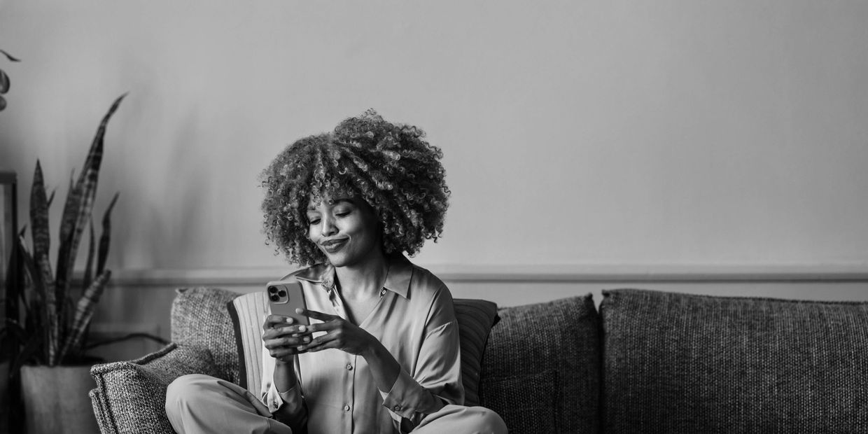 Woman with curly hair sitting on couch, smiling at her phone.