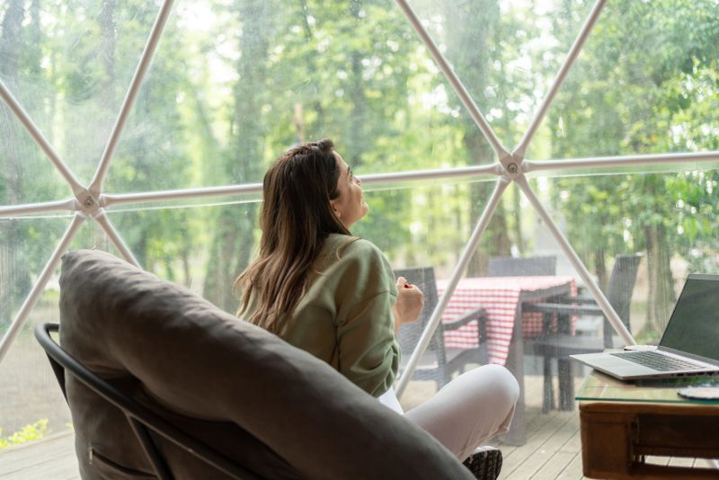 Peaceful Woman Sitting On Sofa At Glamping Camping Hotel Looking At Forest Landscape