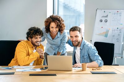 Three colleagues happily collaborate around a laptop in an office.