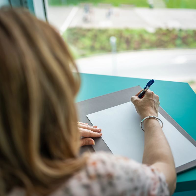 Close-up of a woman writing from behind and next to a huge window overlooking the outside