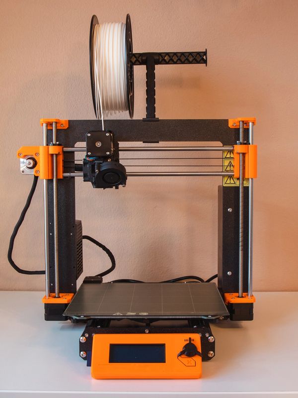 Compact 3D printer with orange accents and white filament spool on a clean white table.