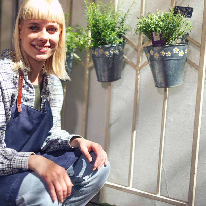 Portrait of a smiling woman sitting at the entrance to a flower shop, there are hanging pots with flowers on the wall
