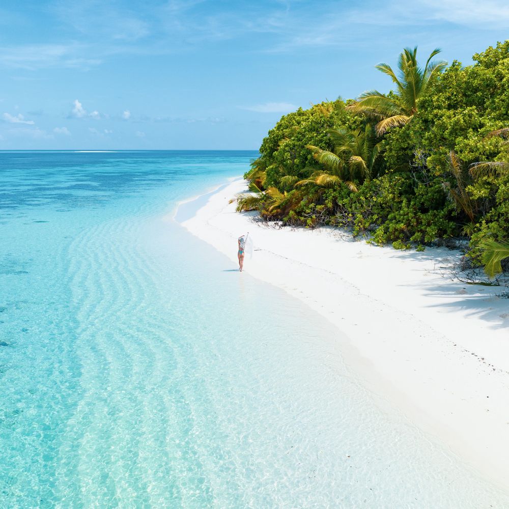 A woman walks along a pristine white beach with turquoise water and lush greenery.