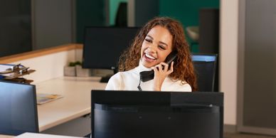 A lady sitting behind a computer monitor, talking on the phone.