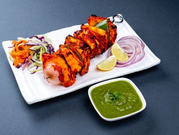 Grilled paneer tikka with green chutney and sliced vegetables on a white plate.