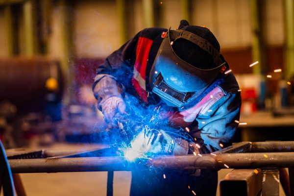 A welder in protective gear sparks while welding metal indoors.