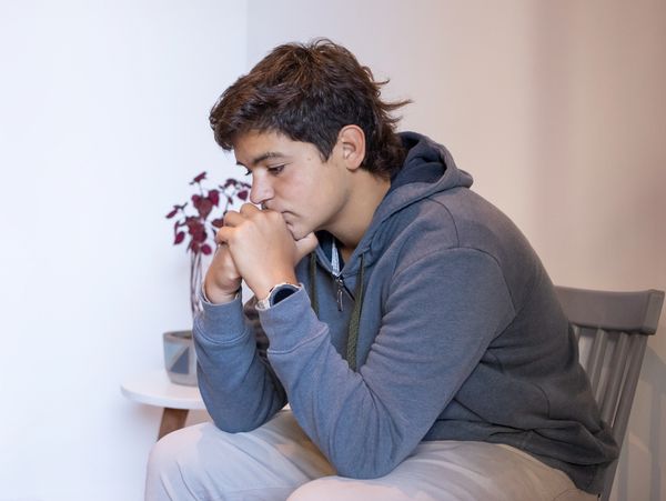 A young man in a hoodie sitting thoughtfully with clasped hands.