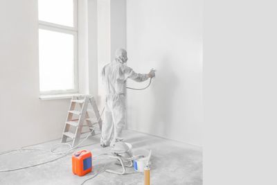 Person in protective suit spray painting a white wall indoors.