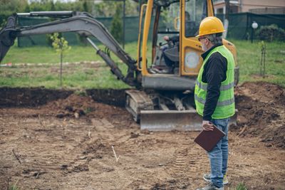 Construction worker in safety gear inspecting a site with an excavator.