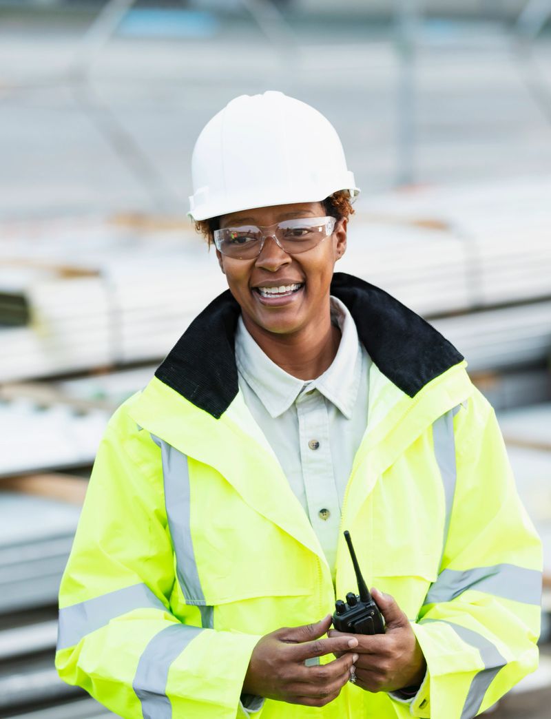 An African-American woman working at a construction site, wearing a hard hat, safety goggles and reflective clothing. She is smiling at the camera, standing in front of stacks of construction material, holding a walkie-talkie. She is a mature woman in her
