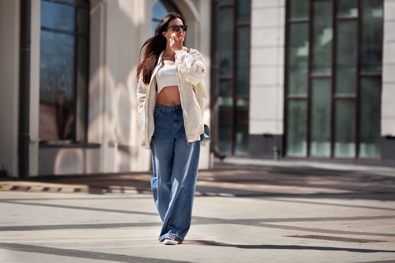 Fashionable woman wearing trendy white bomber jacket, CROP TOP, wide jeans walks on urban city street. Trends clothing of spring and summer.
