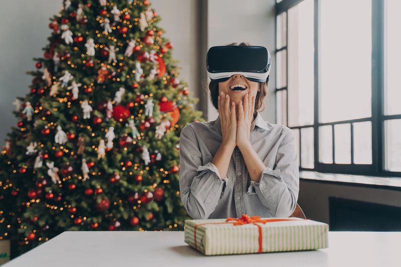 Christmas and future technology concept. Amazed excited young woman wearing virtual reality goggles sitting against xmas tree with gift box at home, astonished female in VR headset playing 3D games
