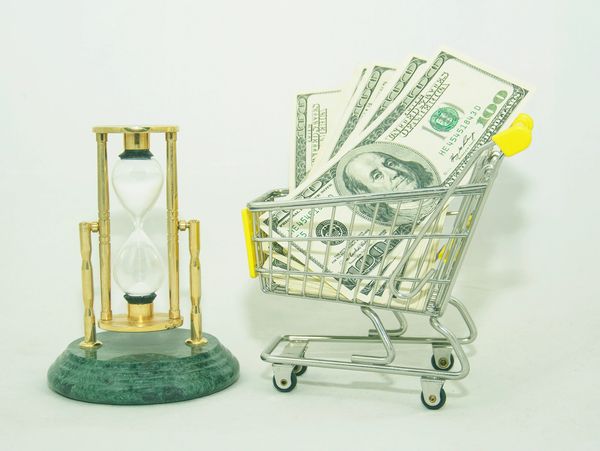 Hourglass next to a shopping cart filled with US dollar bills.