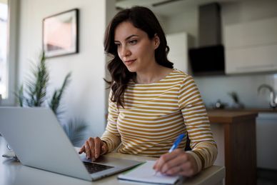 Woman in striped shirt working on laptop and writing notes.
