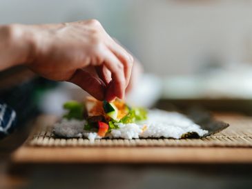 Hand placing cucumber on sushi roll with rice and vegetables.