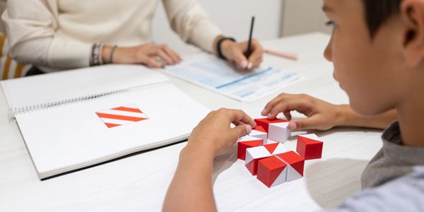 Child solving a red and white puzzle while an adult writes notes.