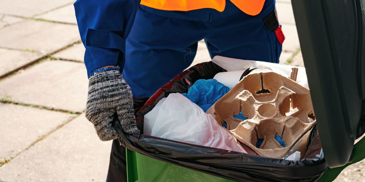 A sanitation worker handling a green trash bin filled with various waste items.