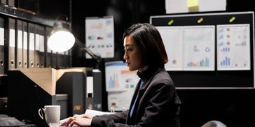 Woman working late on a laptop in a dimly lit office with charts on the wall.