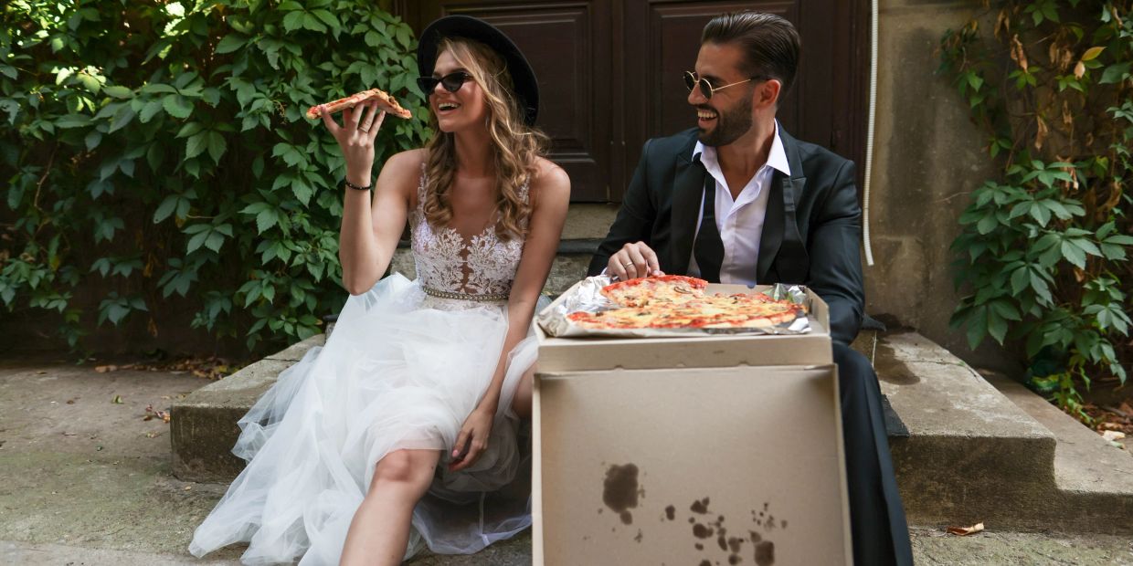 Bride and groom eating pizza outdoors, smiling.