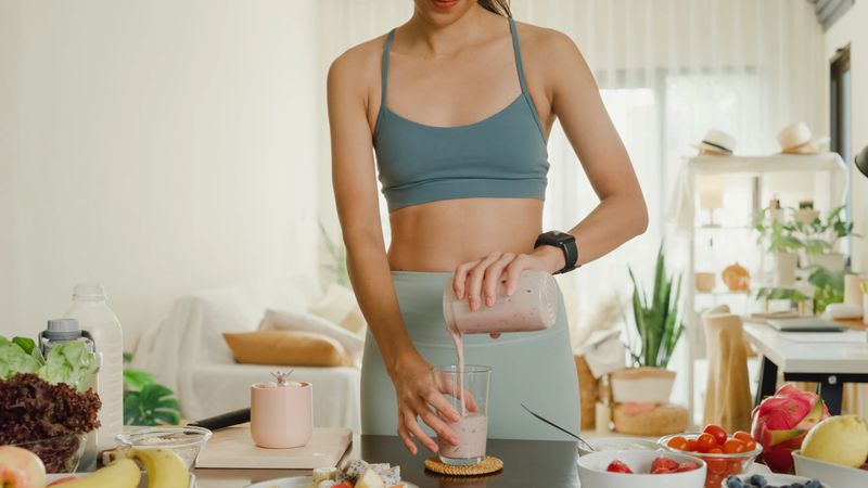 Young Asian athletic woman using blender and pouring smoothie to glass at home. Healthy food concept.