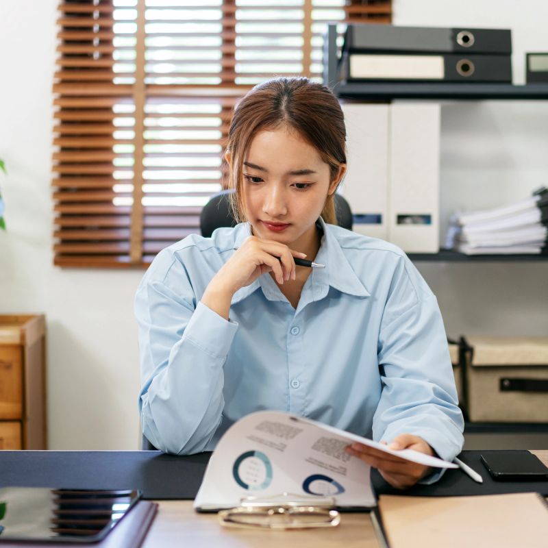 Businesswoman is thoughtful about accounting of business while reading information of new business project in document and working on laptop at modern office.